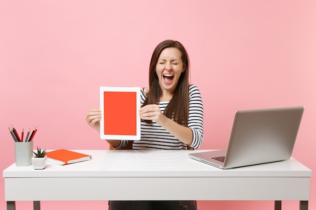 Overjoyed woman with closed eyes hold tablet computer with blank empty screen, sit work at white desk with contemporary pc laptop isolated on pink background. Achievement business career. Copy space.