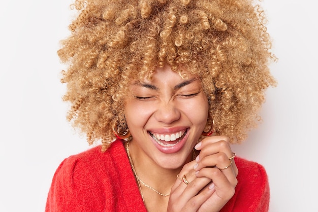 Overjoyed woman with curly hair keeps hands together keeps eyes closed giggles happily dressed in red jumper feels happy isolated over white background. Positive optimistic female being in good mood