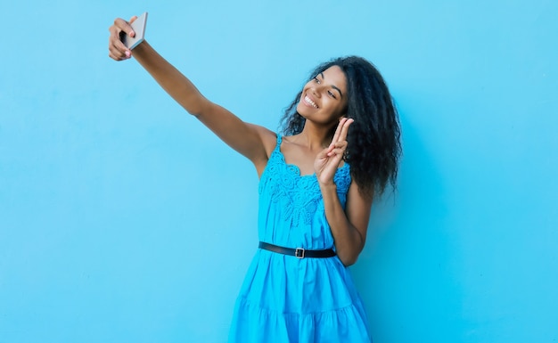 Overjoyed young African ethnic lady is laughing sincerely while holding a smartphone in her stretched arms and taking a selfie