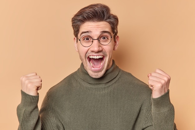 Overjoyed young handsome man exclaims loudly keeps mouth opened
clenches fists rejoices success wears round spectacles casual
sweater isolated over beige background. people and triumph
concept