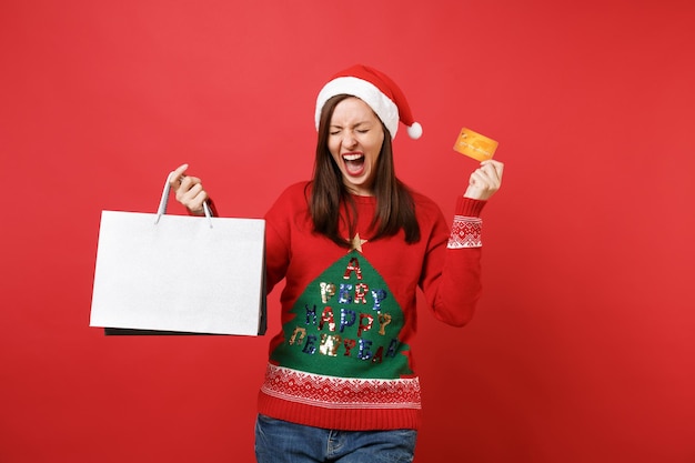 눈을 감고 비명을 지르는 어린 산타 소녀는 빨간색 배경에서 격리된 쇼핑 후 구매한 신용 카드 패키지 가방을 들고 기뻐합니다. 새해 복 많이 받으세요 2019 축 하 휴일 파티 개념입니다.