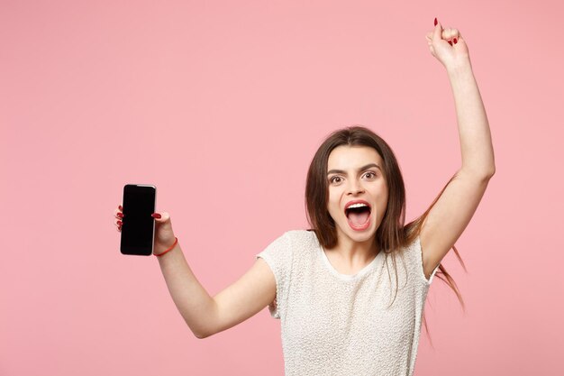 Overjoyed young woman in casual light clothes posing isolated on pastel pink background. People lifestyle concept. Mock up copy space. Hold mobile phone with blank empty screen, doing winner gesture.