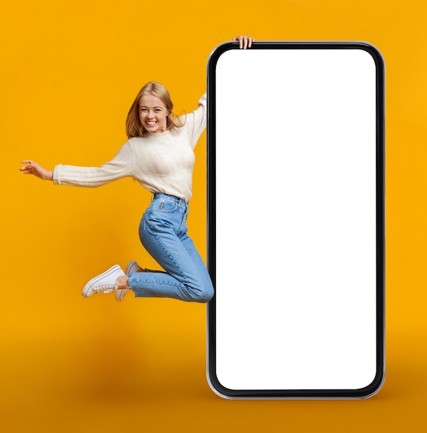 Обрадованная молодая женщина прыгает с огромным пустым смартфоном на желтом фоне