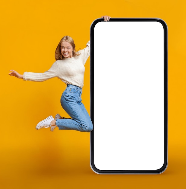 黄色の背景の上に巨大な空白のスマート フォンでジャンプ大喜びの若い女性