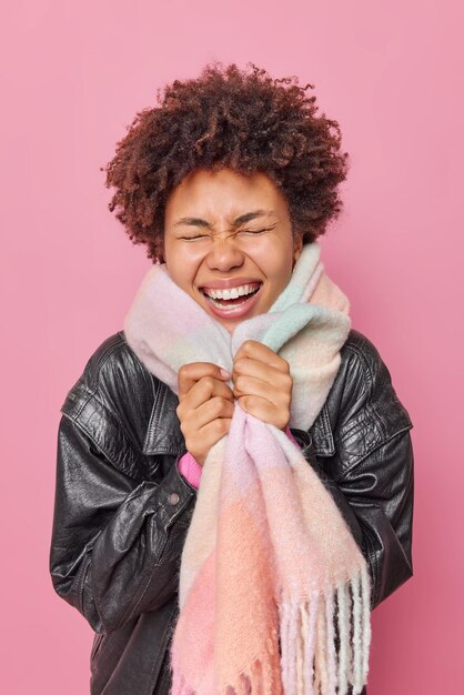 Overjoyed young woman laughs out shows white teeth feels very happy to buy new clothes wears black leather jacket and warm scarf around neck expresses happiness isolated over pink background.