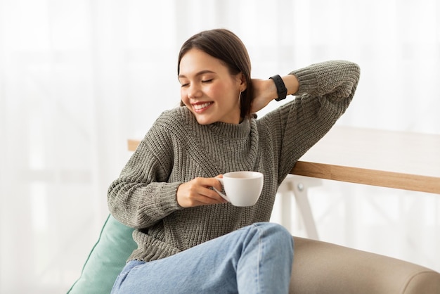 写真 幸せな若い女性が自宅のソファでコーヒーを飲みながらリラックスしています