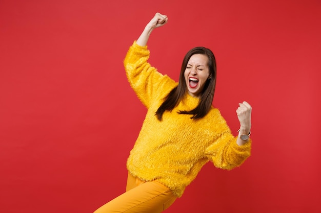 Overjoyed young woman in yellow fur sweater with closed eyes doing winner gesture, screaming isolated on red wall background in studio. People sincere emotions, lifestyle concept. Mock up copy space.