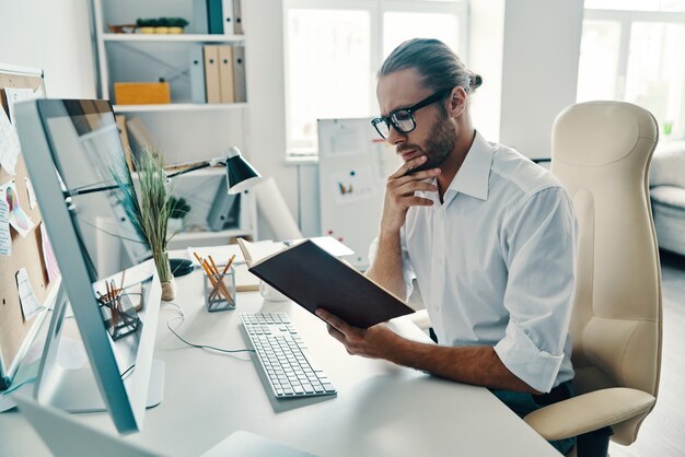 Overthinking. Good looking young man in shirt reading his note pad while sitting in the office