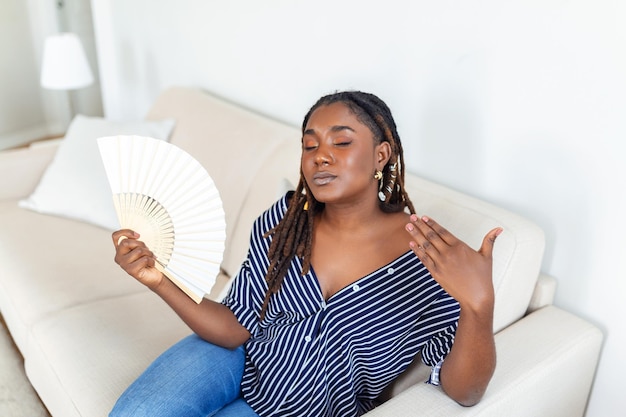 Oververhitte Afrikaanse vrouw zit thuis op de bank, voelt warm zwaaiend met de handventilator die afkoelt zwetend meisje ontspant op de bank in de woonkamer houd wankelend last van hitte geen airconditioningsysteem