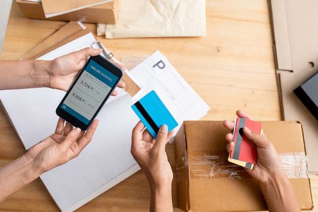 Photo overview of hands of two female managers holding smartphone with online banking operation and plastic cards over workplace