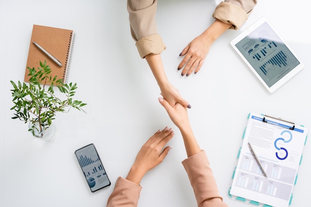 Overview of hands of two young successful businesswomen handshaking over desk among financial information on tablet screen and paper