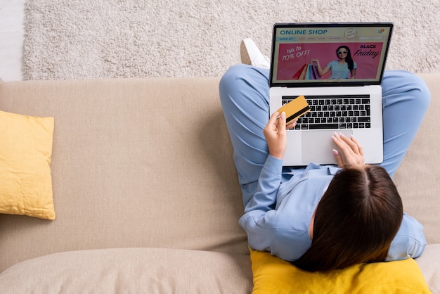 Overview of young brunette female in blue pajamas sitting on couch in living-room and going to pay by card for goods from online shop