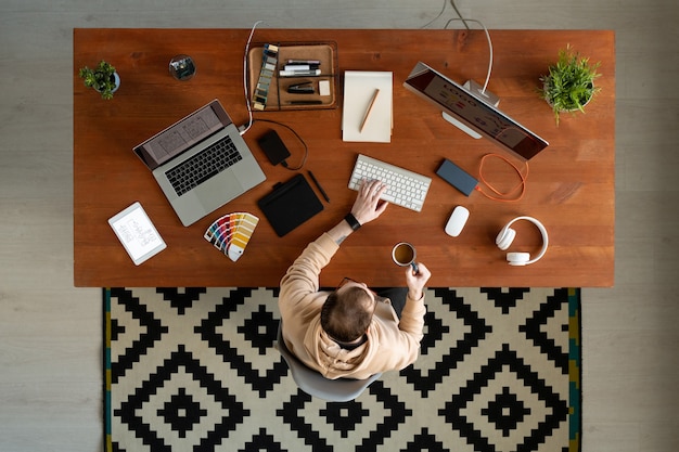 Overview of young contemporary male freelance designer in casualwear pressing buttons of computer keypad while having coffee by table