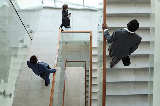 Overview of young intercultural business people walking upstairs and downstairs inside contemporary office building