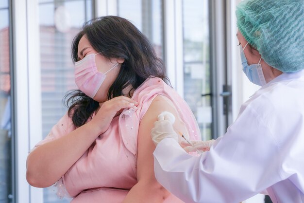 과체중 아시아 여성 환자가 Covid 19에 대한 주사 예방 접종을 위해 간호사에게 팔을 제공합니다.
