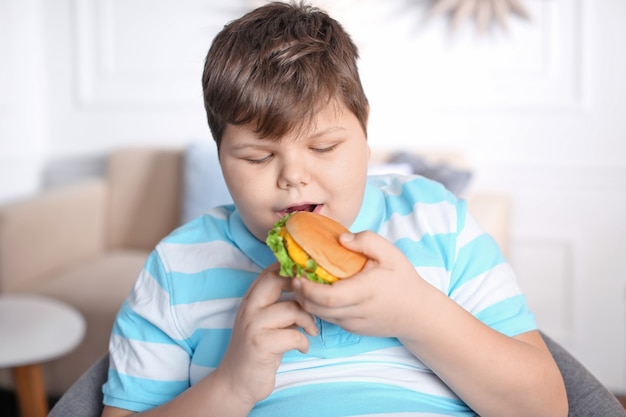 Ragazzo in sovrappeso che mangia hamburger al chiuso