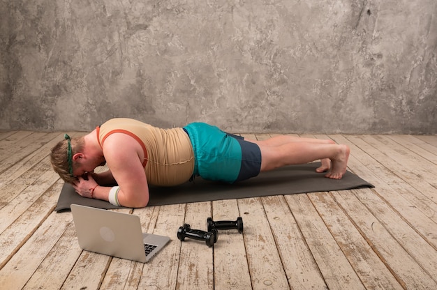 Uomo in sovrappeso che si esercita, facendo esercizi di stretching sul tappetino yoga, guardando video di fitness online sul laptop a casa.