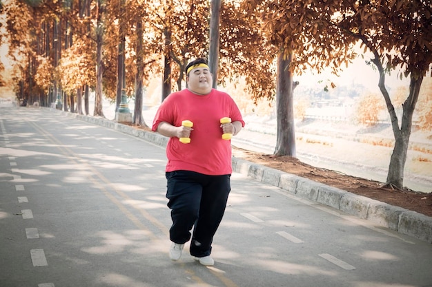 Photo overweight man holding dumbbells while running on road in park