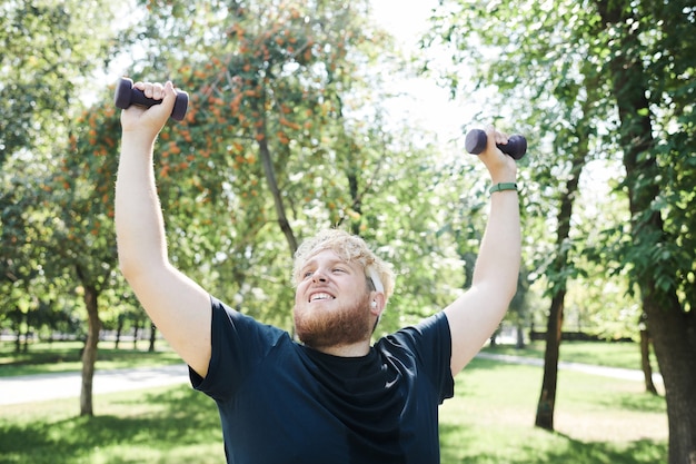 Мужчина в спортивной одежде с избыточным весом делает упражнения с гантелями на открытом воздухе
