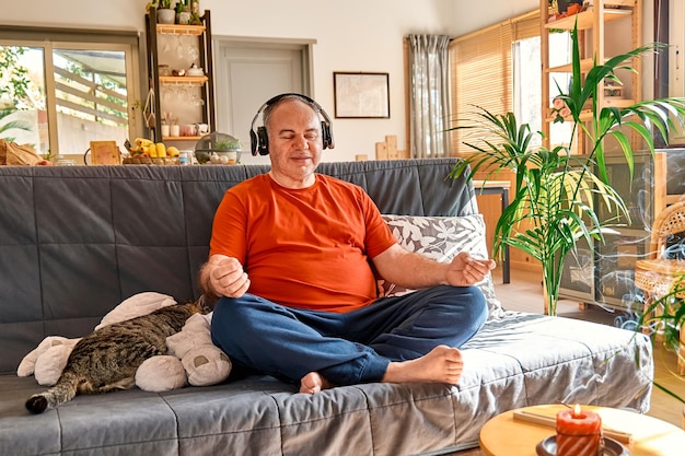 Мужчина с избыточным весом в беспроводных наушниках практикует йогу и медитацию под руководством дома, сидя в позе лотоса на диване и слушая расслабляющую музыку. Концепция осознанной медитации