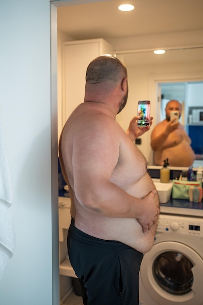 사진 집의 욕실에서 거울을 보고 스마트폰으로 셀카를 찍는 벌거벗은 몸통을 가진 과체중 남성