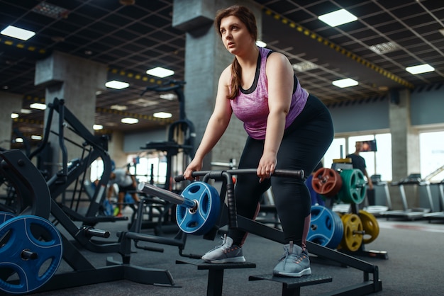 Overweight woman doing exercise with bar in gym, active training.