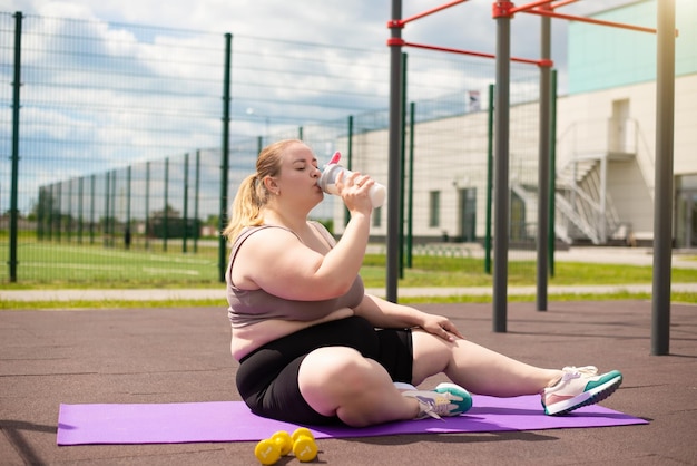 Женщина с избыточным весом пьет протеиновый коктейль из бутылки, сидя на коврике во время тренировки.