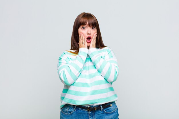 Overweight woman feeling shocked and scared, looking terrified with open mouth and hands on cheeks