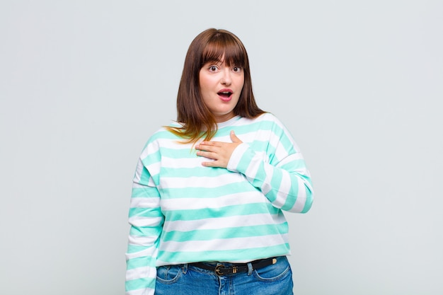 Overweight woman feeling shocked and surprised, smiling, taking hand to heart, happy to be the one or showing gratitude