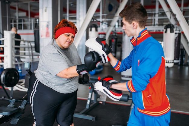 Overweight woman in gloves boxing with instructor in gym. 