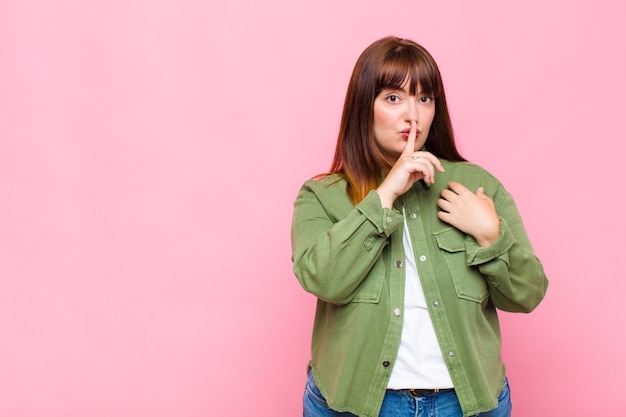 Overweight woman looking serious and cross with finger pressed to lips demanding silence or quiet, keeping a secret