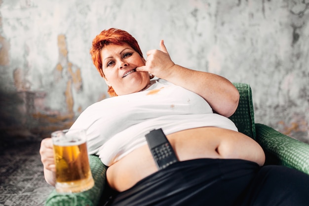 Женщина с избыточным весом сидит в кресле и пьет пиво, страдает булимией, ожирением. Нездоровый образ жизни, толстая самка