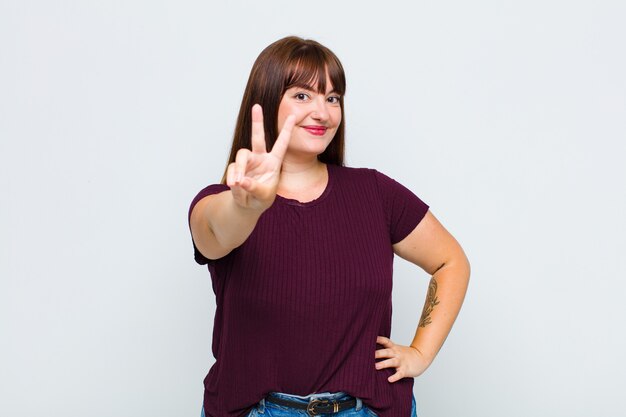 Overweight woman smiling and looking happy, carefree and positive, gesturing victory or peace with one hand