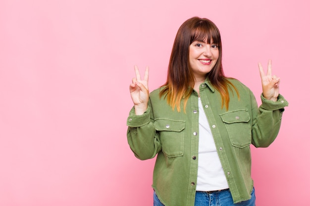 Overweight woman smiling and looking happy, friendly and satisfied, gesturing victory or peace with both hands