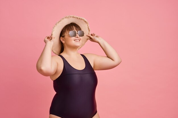 Overweight woman in swimsuit prepares for summer vacations, body positive, pink wall