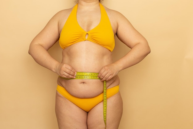 Overweight woman in yellow swimsuit measure waist circumference with measuring tape beige background Slimming and body control struggle with obesity Plus size people and body positive concept