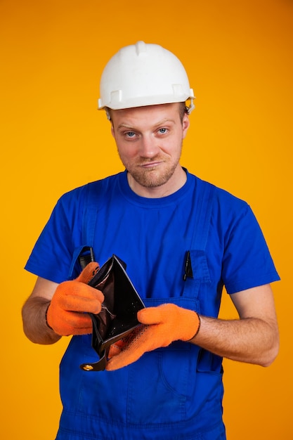 Il lavoratore in sovrappeso in un casco protettivo mostra un portafoglio vuoto. recessione nell'economia. un uomo in tuta senza soldi. recessione e crisi economica, disoccupazione