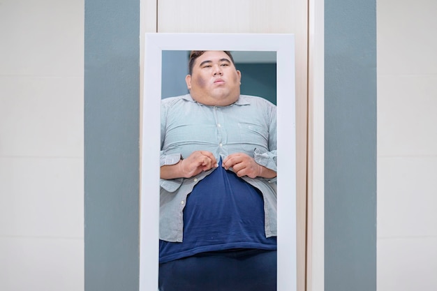 Overweight young Asian male looking at mirror with tight shirt