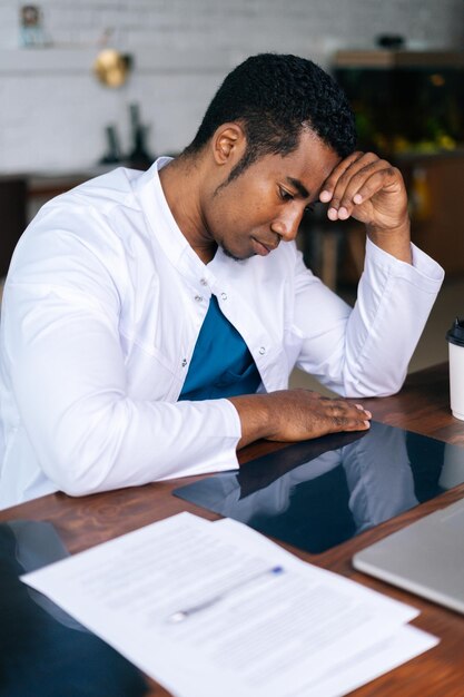 Overwerkte jonge Afro-Amerikaanse zwarte mannelijke arts in witte jas koffie drinken tijdens het werken typen op laptopcomputer zittend op moderne kantoor Concept van geneeskunde en gezondheidszorg