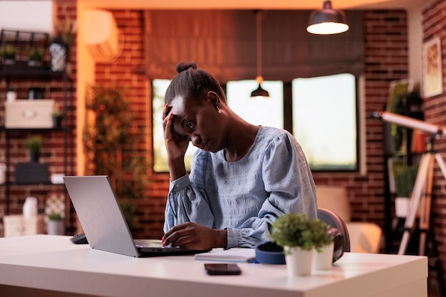 Overwerkte jonge vrouwelijke freelancer die op laptop werkt in een modern thuiskantoor, burn-out en werklastconcept. gestresste en uitgeputte afro-amerikaanse zakenvrouw met hoofdpijn