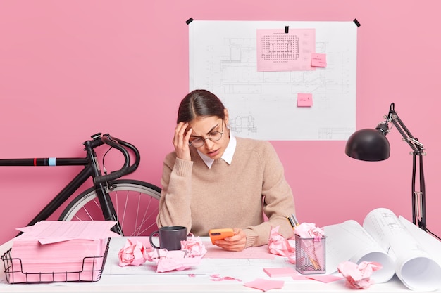 Overwerkte vermoeide vrouwelijke architect gericht op smartphone heeft veel werk te doen werken aan architecturaal project maakt schetsen trekt schetsen poses in coworking-ruimte tegen roze muur. Kantoormedewerker