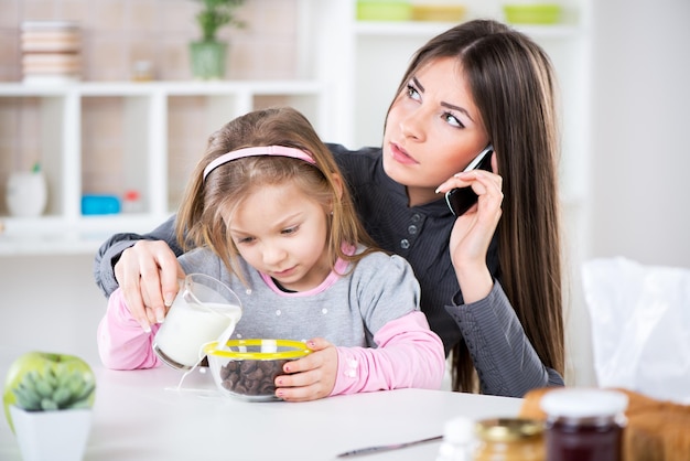 Overwerkte zakenvrouw en haar dochtertje in de ochtend. Overwerkte moeder telefoneert voordat ze naar haar werk gaat. Voorbereiding van granen met melk voor haar dochter en het morsen van melk naast de kom.