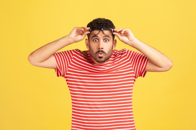 Overwhelmed bemused man with beard in striped t-shirt raising eyeglasses and looking at camera with big eyes and shocked surprised expression. Indoor studio shot isolated on yellow background