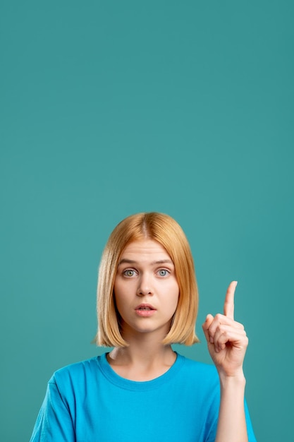 Overwhelmed woman Exciting opportunity Portrait of inspired blonde lady in blue tshirt pointing up at copy space isolated on teal commercial background Surprising idea