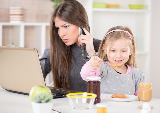 Donna d'affari oberata di lavoro e la sua piccola figlia al mattino. la mamma leggeva la posta e telefonava prima di andare al lavoro. figlia che spalma il burro di arachidi sul pane