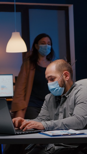 Overworked businessman with face mask against covid working in startup office checking management st...