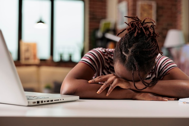 Libero professionista oberato di lavoro che dorme sul tavolo durante il giorno, lavoratore remoto stressato assonnato, esaurimento professionale. studente stanco sdraiato sulla scrivania, impiegato afroamericano esausto che ha una pausa sul posto di lavoro