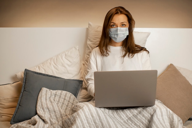 Overworked girl in face mask works at laptop in bed,