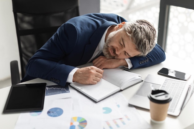 Uomo d'affari di mezza età oberato di lavoro che fa un pisolino sul posto di lavoro in ufficio