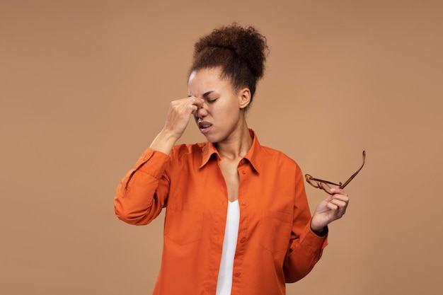 Overworked multitasking woman holds glasses feels headache after hardworking day beige background